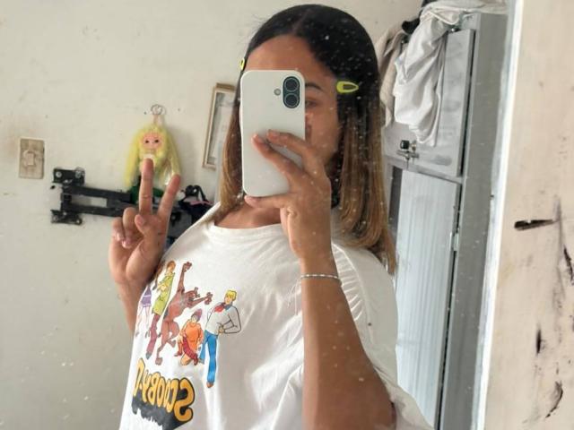 KendallKiss tirando uma selfie no espelho, exibindo sua camisa branca com uma estampa colorida de desenho animado, fazendo um sinal de paz com a mão direita, com o celular cobrindo seu rosto..