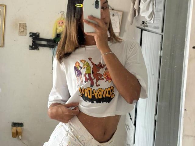 KendallKiss, com pele bronzeada, cabelo castanho e óculos, vestindo uma camiseta branca com personagens de desenhos animados e a frase "900+000%", levantando a camiseta para mostrar sua barriga plana e calças brancas..