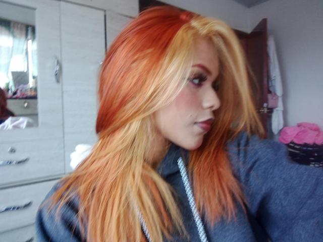 IvonneKross with long, orange and yellow streaked hair, wearing a blue shirt, has a pouty expression, and is looking to the side. Her tits are slightly visible.
