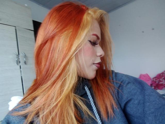 IvonneKross with orange and yellow hair, wearing a blue shirt, has a nose piercing, and is looking down with her eyes closed. Her tits are small and she's cute.
