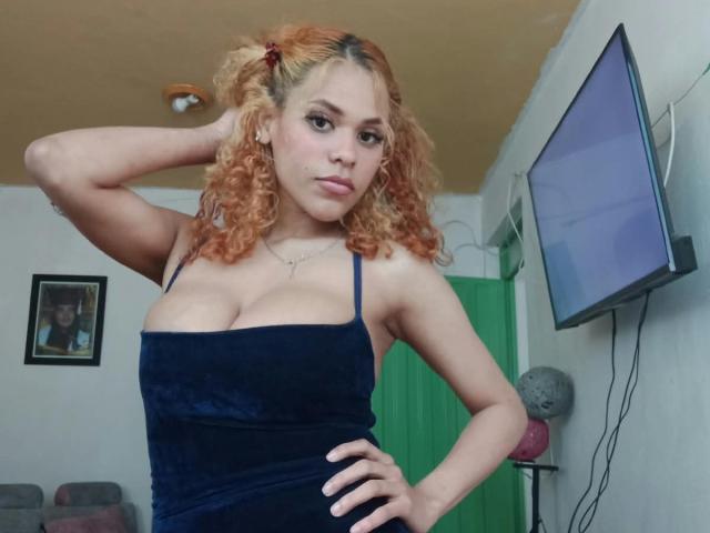 IvonneKross with curly orange hair, wearing a tight blue velvet dress, showing her big tits, hand on hip, looking at the camera, in a room with a flat screen.