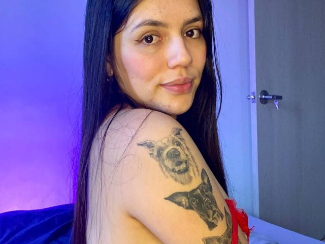 SophiaMarx with long black hair, smiling, looking over shoulder, showing a tattoo of a wolf and bat on her right shoulder, topless with red bra straps.