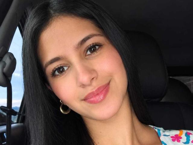 Amily Preston tem cabelo preto, olhos castanhos, batom rosa e um leve sorriso. Ela está usando pequenos brincos de ouro e uma blusa colorida. Ela está sentada em um carro..