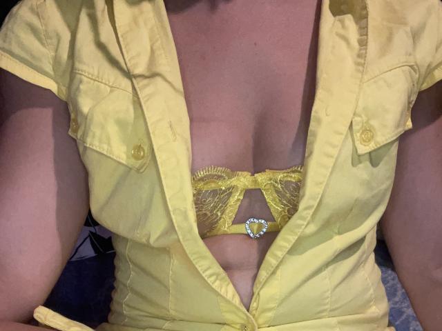 O sutiã amarelo da Lunevia, com renda e uma joia em formato de coração, é visível sob sua camisa amarela desabotoada, revelando seus seios. Ela tem um tom de pele claro e está sentada..