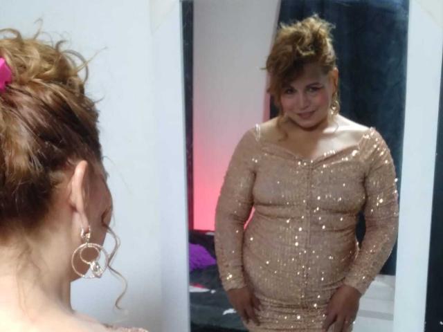 Kazsandra mit lockigem, braunem Haar, das mit einem rosa Haargummi zusammengebunden ist, trägt ein glitzerndes, eng anliegendes, beiges Kleid und steht vor einem Spiegel, wobei sie ihre Brüste und ihren Hintern zeigt..