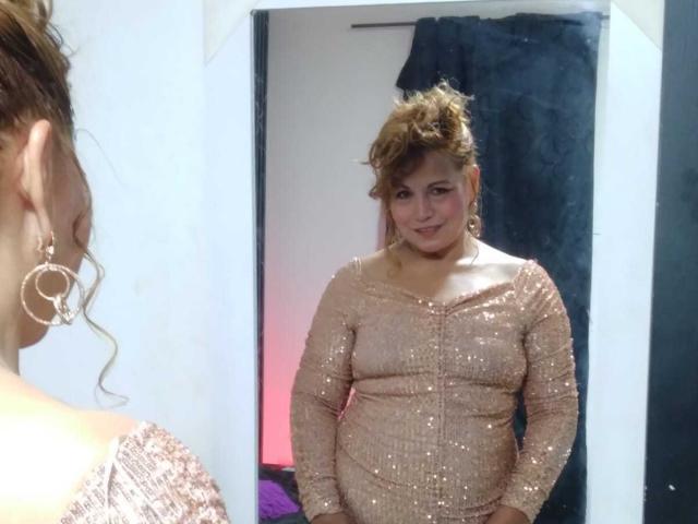 Kazsandra with curly brown hair stands in front of a mirror, wearing a shiny, pink, long-sleeve dress, showing her curvy, full-figured body and smiling at herself.