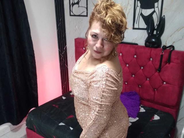 Kazsandra with curly blonde hair, wearing a sparkly off-shoulder dress, sitting on a red tufted bed, looking back with a smile, showing her ass slightly.