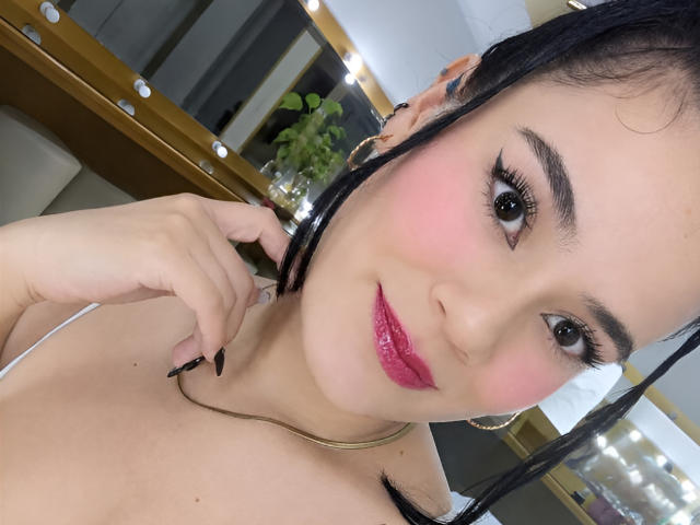 Isabella Voss, com bochechas rosadas, batom rosa brilhante, delineador preto, brincos de argola que tocam o pescoço, esmalte preto nas unhas e cabelo preto brilhante, em uma selfie no espelho..