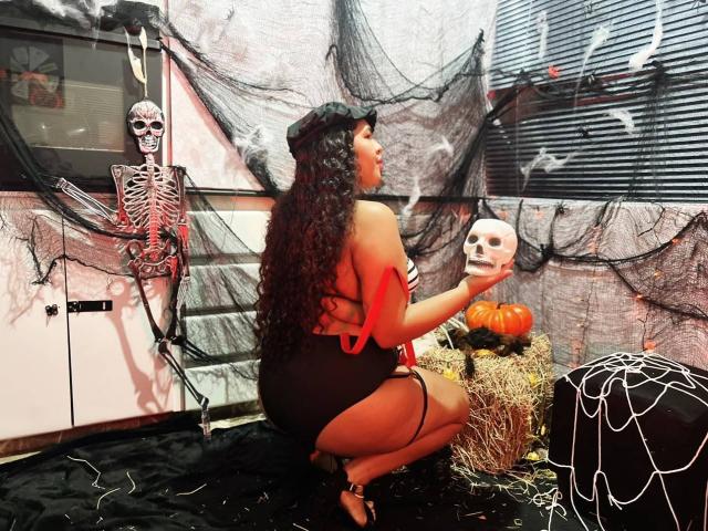 AshleyJenner, une femme aux formes généreuses avec de longs cheveux noirs, est accroupie, tenant un crâne, et porte une robe noire et rouge, des talons noirs et un chapeau noir, dans un décor d'Halloween effrayant..