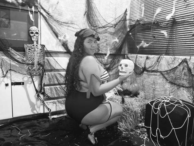 AshleyJenner, pulpeuse avec de grosses fesses, tenant un crâne, portant une casquette et un haut rayé, accroupie près d'un squelette et d'une citrouille, en noir et blanc, avec de longs cheveux bouclés, une ambiance Halloween effrayante..