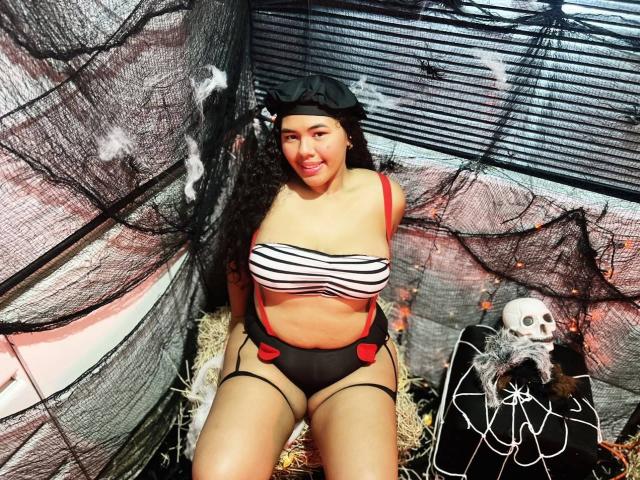 AshleyJenner with curly black hair, wearing a black cap, striped tube top, black and red panties with suspenders, and a skull decoration on the right. Boobs, ass, dark skin.