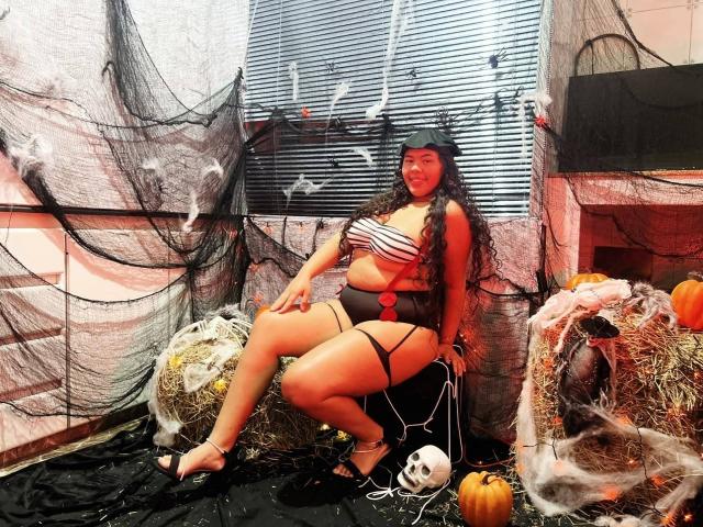 AshleyJenner, curvy with big boobs and a round ass, sits in a Halloween setup wearing a striped bra and black garter, with long hair, a black headband, and high heels.