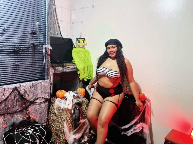 AshleyJenner, avec de longs cheveux bouclés, portant un bonnet noir, un soutien-gorge rayé, une jarretière noire et un short noir, assise à côté d'un costume de hibou vert, entourée de décorations d'Halloween..