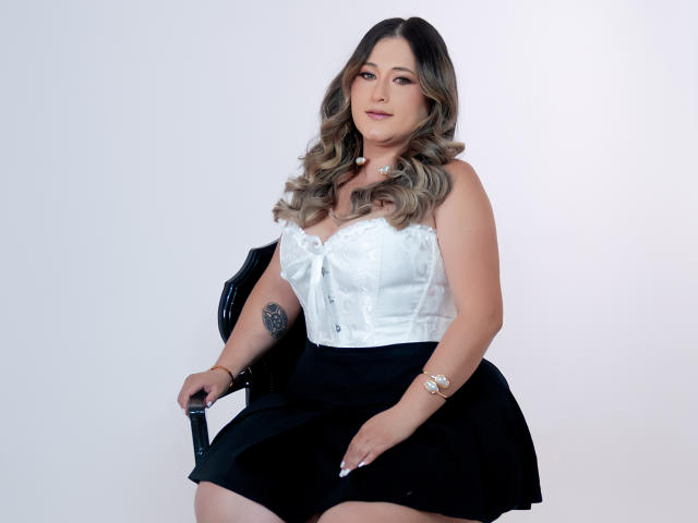 BeccaSwan, con cabello rubio ondulado, corpiño blanco de encaje, falda negra, tatuaje en el brazo izquierdo, sentada en una silla negra, mirando a la cámara, con un brazalete, sexy, curvilínea..