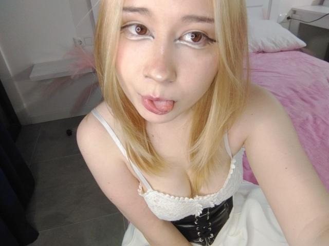 AnnieLeonhart69 with blonde hair, sticking out her tongue, wearing a white lace top with black corset, showing her small tits, and pale skin, in a bedroom.