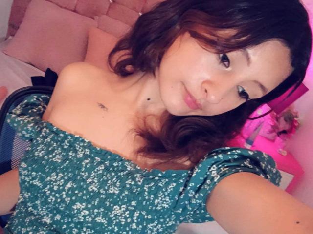 ValentinaRoseX with wavy brown hair, green floral off-shoulder top, small tattoo near collarbone, cute smile, pink bedroom, pink headboard, pink lamp, soft pink lighting.