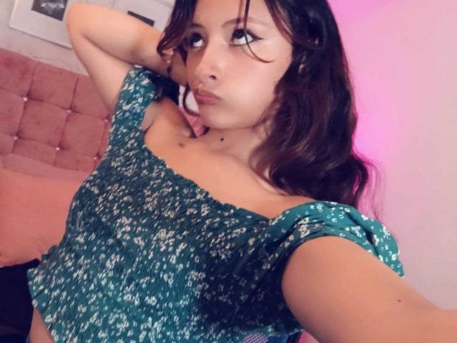 ValentinaRoseX with pouty lips, dark wavy hair, green off-shoulder top with white patterns, one arm behind head, small mole above her tits, pink and white room.