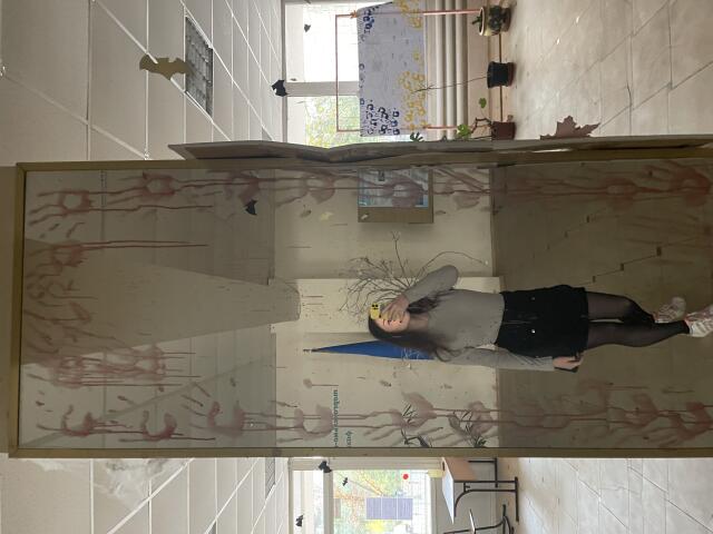 Ellaria stands in a grungy hallway with a dirty glass panel in front of her, wearing a gray top, black skirt, and black tights, with her ass visible.