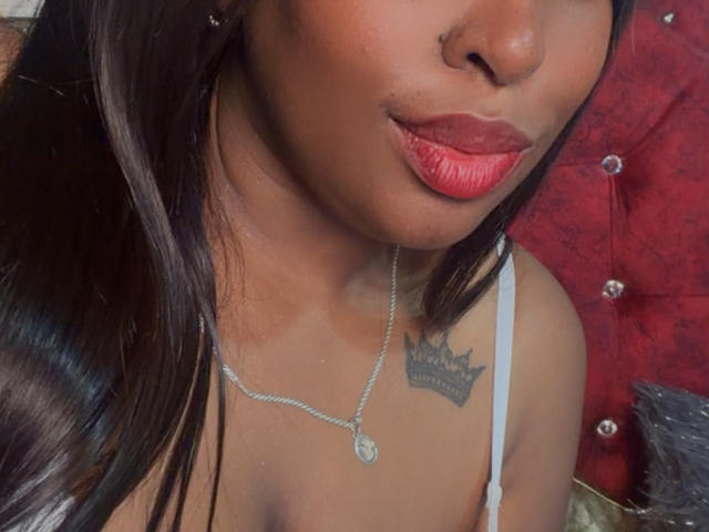 NatyLm has dark brown skin, red lipstick, straight black hair, a silver necklace, and a crown tattoo on her right shoulder, wearing a white strap top.
