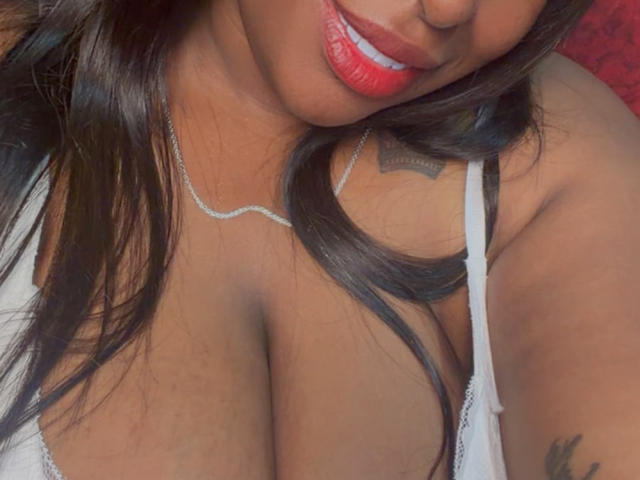 NatyLm's busty chest with large tits, wearing a white lace top, has red lipstick and a silver necklace, with long dark hair and a tattoo on her right shoulder.