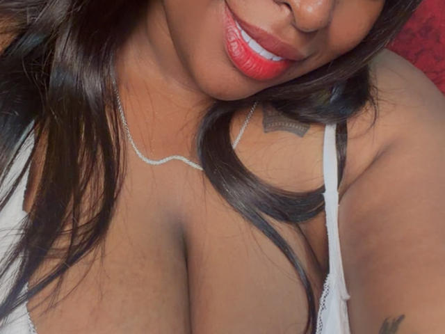 NatyLm with red lipstick, brown hair, and a white bra, showing her large tits and a tattoo on her right shoulder. She's wearing a silver necklace.