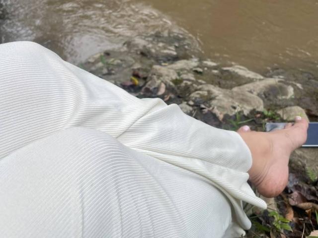 As nádegas e pernas de NahomyStone, vestindo calças brancas texturizadas, com os pés à mostra, um deles levemente curvado, ao lado de uma pedra lamacenta e água, com um smartphone na grama..