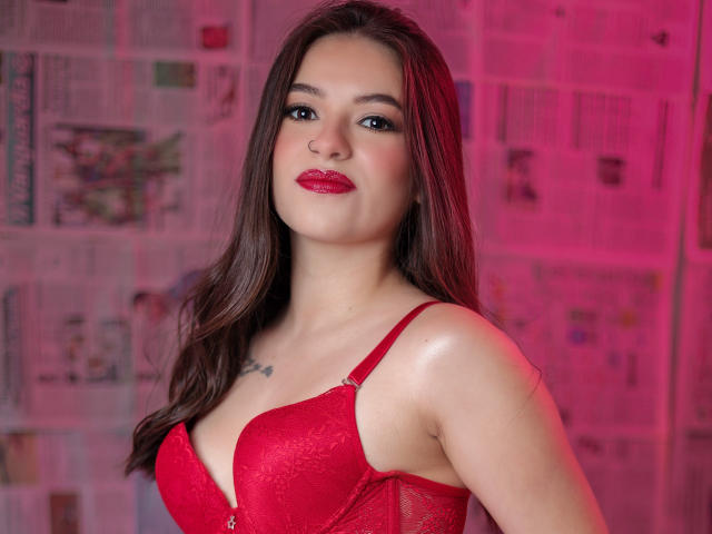 PaolaYKatty with red lace bra, medium tits, long brown hair, red lipstick, slight smile, tattoo above chest, pink light, newspaper background.