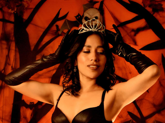 BrianaHarpers with dark curly hair, wearing a black leather bra, black gloves, and a sparkly tiara, smiling with eyes closed, showing her tits and cute face.