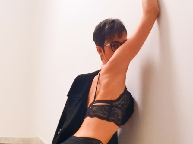 FrancaisePhoenix with short black hair, glasses, black lace bra, and black pants, leaning against a white wall, arm raised, black jacket falling off shoulders, showing her tits.