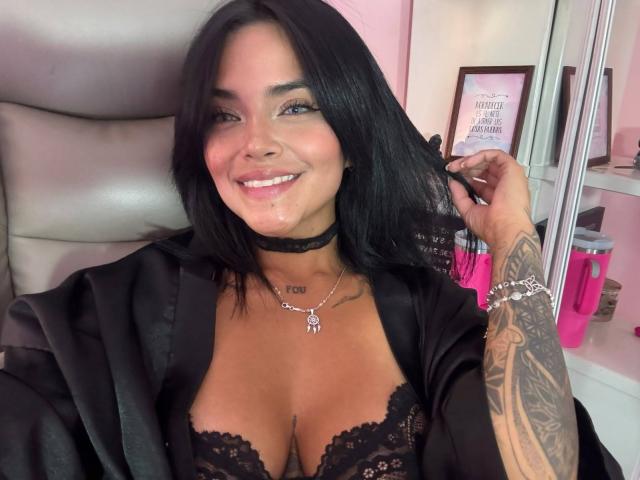 MajoJones has black hair, blue eyes, and a tattooed arm. She's smiling, wearing a black choker, lace bra, and open robe, showing her boobs. She's holding her hair.