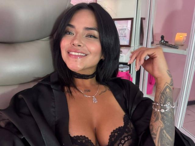 MajoJones mit schwarzem Spitzen-BH, der ihre Brüste zeigt, lächelnd, Sperma im Gesicht, schwarzer Choker, Tattoos am rechten Arm, sitzend auf einem Stuhl, trägt einen schwarzen Umhang..
