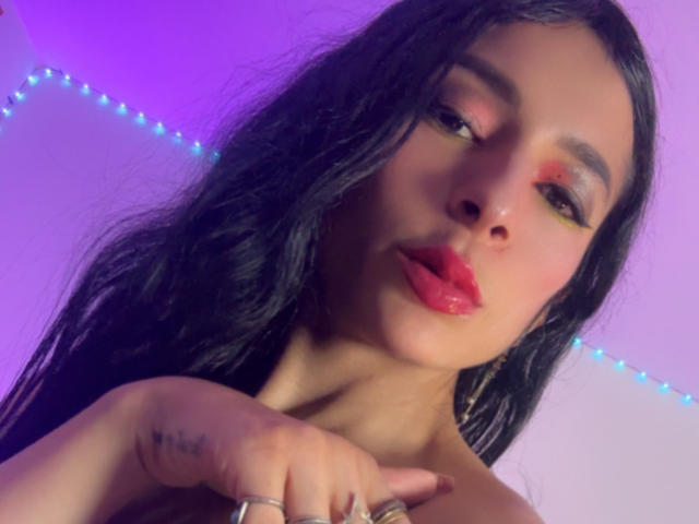 Ariel Nitzche, met lang zwart haar, rode lippenstift, gele oogschaduw en tatoeages op haar vingers, wijst naar de camera. In de achtergrond zijn paarse en blauwe LED-stripverlichting te zien..