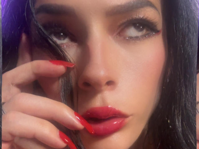 Le visage d'ArielNitzche, avec ses cheveux foncés, ses lèvres rouges et ses longs ongles rouges, touchant sa joue avec une expression sensuelle, des yeux brillants et des lèvres pulpeuses légèrement entrouvertes..