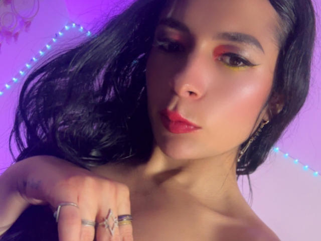Ariel Nitzche, avec ses cheveux noirs et ondulés, son fard à paupières rouge, son crayon à paupières jaune, son rouge à lèvres rouge et ses multiples bagues aux doigts, a une allure sexy et confiante, avec un arrière-plan illuminé de violet..