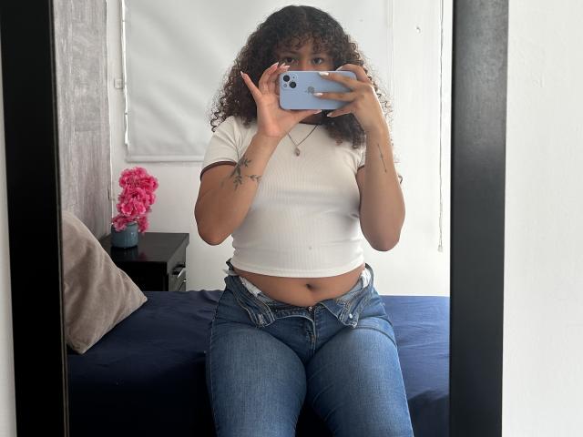 Sofia, passionnée, avec des cheveux bouclés, un débardeur blanc laissant apparaître son ventre, et un jean bleu, tenant un téléphone, se cachant le visage, assise sur un lit, selfie devant un miroir, fleurs roses..