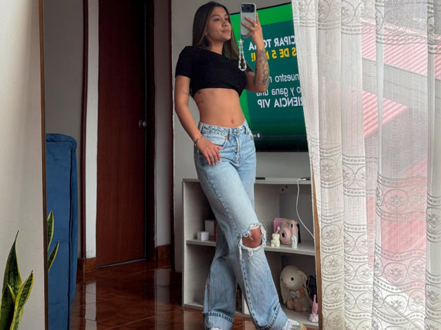 Thalia Ares, con cabello castaño largo, un top corto negro y jeans azules rotos, tomando una selfie frente al espejo, mostrando sus abdominales tonificados y pequeños tatuajes en su brazo derecho..