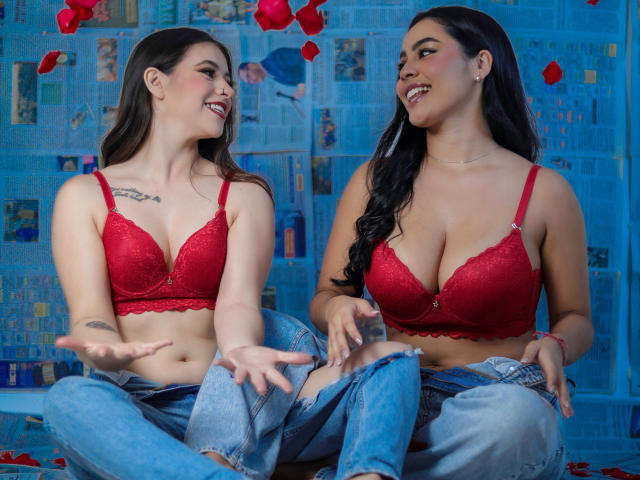 PaolaYKatty and PaolaYKatty, both with red lace bras, showing off their tits, sitting cross-legged, laughing, and gesturing with hands, petals scattered on the floor.