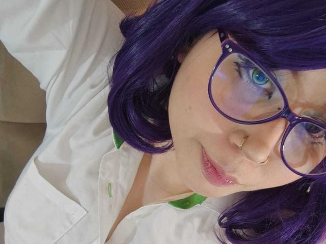 WendyMonet with purple hair, glasses, and nose ring, wearing a white shirt, showing blue eyes and pink lips, holding a green object. Boobs partially visible, slightly open shirt.