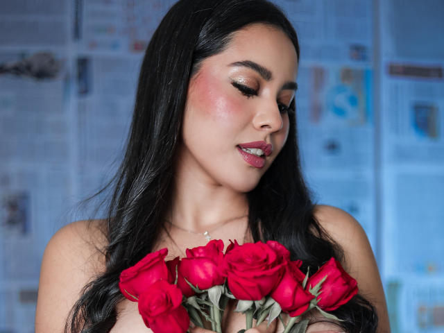 PaolaYKatty, con capelli scuri, fard rosa e rossetto rosso, che tiene rose rosse, con gli occhi chiusi, senza indumenti sopra, mostrando il seno, sfondo sfocato..