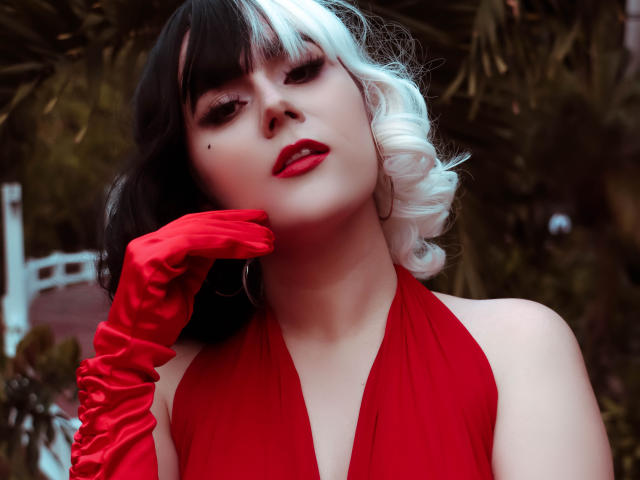 GoddessBar with black and white curly hair, red lipstick, red halter dress, and red satin gloves, touching her cheek, looking seductively at the camera. Boobs visible, ass not.