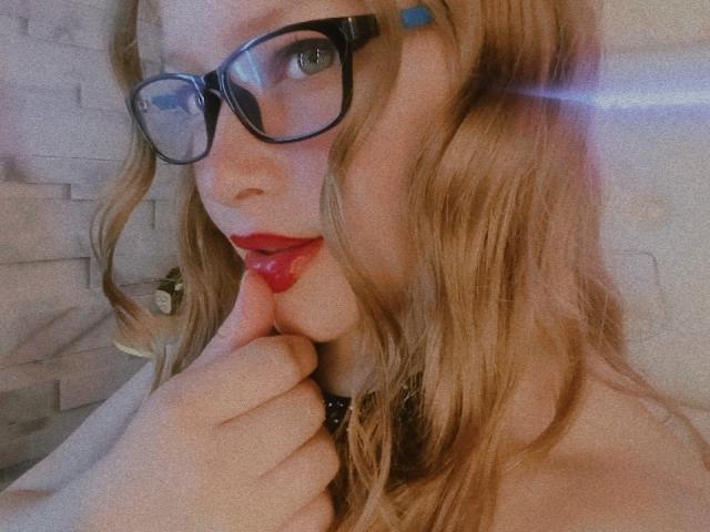 IvyBellmont met een bril, rode lippenstift en blond golvend haar, die haar lip aanraakt met een vinger, draagt een zwarte choker. Haar borsten zijn gedeeltelijk zichtbaar. Wazig lichteffect..