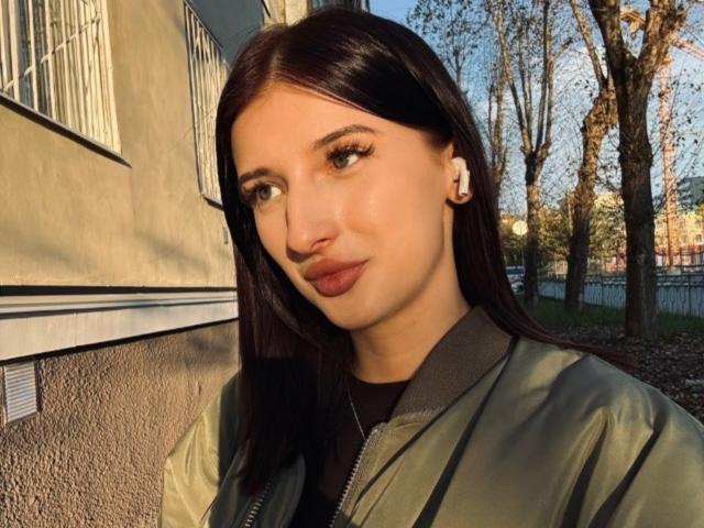 DorianWindley a les cheveux longs et bruns foncés, des yeux verts, et porte une veste verte avec un t-shirt noir. Elle a un petit nez et regarde sur le côté..