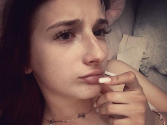 Dorian Windley, mit heller Haut, braunen Haaren und langen Wimpern, beißt an ihrem mit rosa Nagellack lackierten Finger, trägt eine zarte Halskette und wirkt leicht verärgert. Ihre Brüste sind nicht sichtbar..