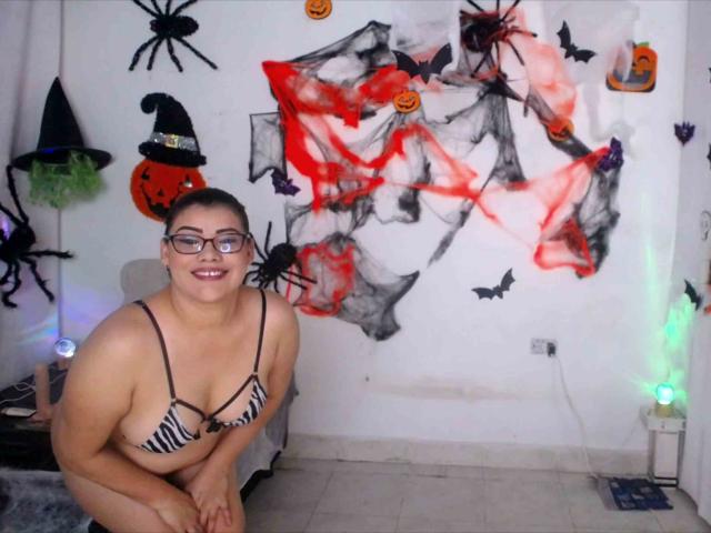 ValeriaLoveer, una donna formosa con gli occhiali, in costume da bagno a righe zebrate, sorridente, con un seno prosperoso e un viso carino, è inginocchiata in una stanza decorata per Halloween, con decorazioni spaventose..