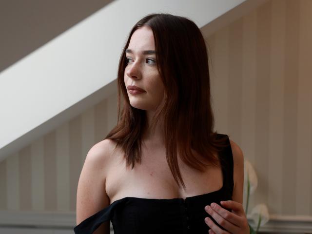 KiaraTease with medium brown hair, looking to the side, has a black dress slipping off her tits, exposing her small, pale-ass. She's standing indoors, looking pensive.