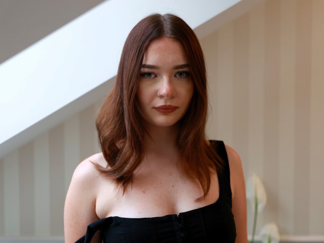 KiaraTease has medium-length brown hair, fair skin, and small, freckled tits in a black, strap-down top. She looks serious with a neutral expression.