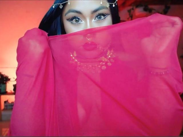 Estefaniaa69 with dark blue hair, wearing a sheer pink dupatta covering her tits and face, showing her green eyes and red lipstick, adorned with a gold necklace and headpiece.