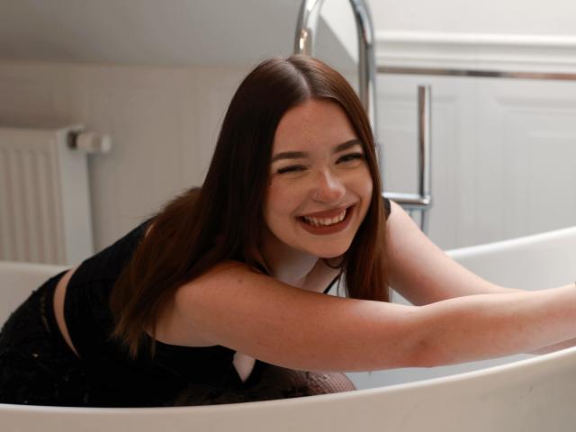 KiaraTease, a cute girl with brown hair, is leaning in a white bathtub, smiling with her teeth, wearing a black top, showing her small tits, and a happy expression.