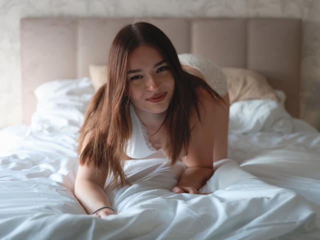 KiaraTease is on all fours on a bed, wearing a white top, her ass is raised, she has long brown hair, and she's smiling with red lipstick.