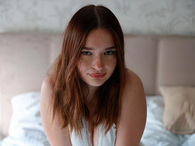 KiaraTease with red hair, freckles, and nose ring, looking up with a slight smile, wearing a white tank top, sitting on a bed with a beige headboard.